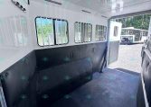Interior of a white horse trailer with barred windows and a black non-slip floor, open rear door, outdoor trailers visible outside.
