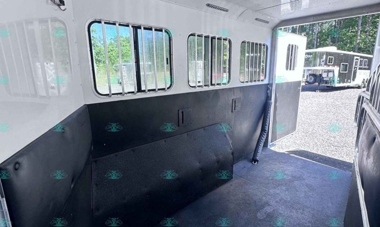 Interior of a white horse trailer with barred windows and a black non-slip floor, open rear door, outdoor trailers visible outside.