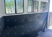 Interior of a horse trailer with barred windows and a dark lower wall, showing a vertical divider and entry at right side, outdoors visible through windows
