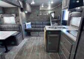 Interior of a modern travel trailer: kitchen with double sink and stove on the right, gray cabinets, and a gray sofa on the left; wood-look flooring.