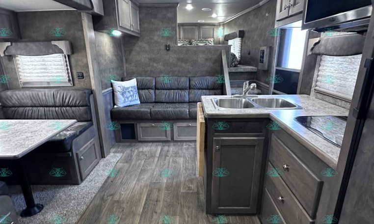 Interior of a modern travel trailer: kitchen with double sink and stove on the right, gray cabinets, and a gray sofa on the left; wood-look flooring.
