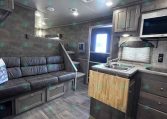 Interior of a modern gray RV living area with a black leather sofa, built-in cabinets, and a kitchen island with a wood cutting board.