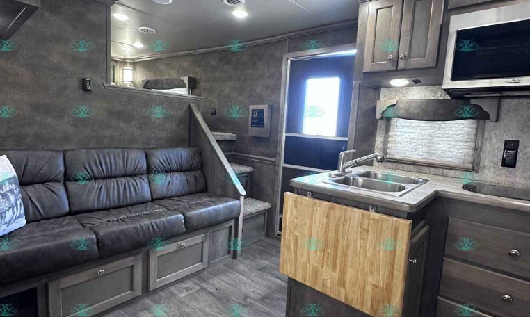 Interior of a modern gray RV living area with a black leather sofa, built-in cabinets, and a kitchen island with a wood cutting board.