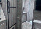 Interior of a gray-toned RV with a tall black-framed refrigerator, wood cabinets, and a doorway to a small bathroom area.