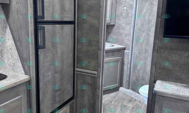 Interior of a gray-toned RV with a tall black-framed refrigerator, wood cabinets, and a doorway to a small bathroom area.