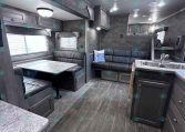 Interior of a modern gray travel trailer: dinette with bench seating, sofa, and kitchen island; overhead cabinets and windows with blinds.