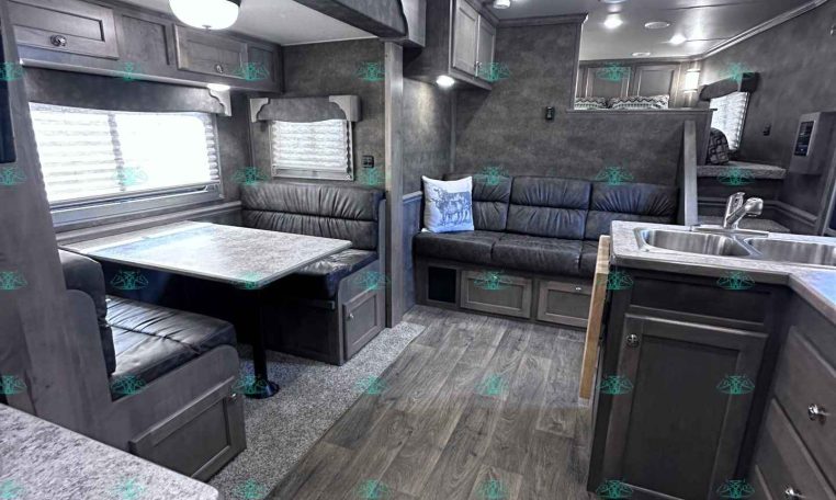 Interior of a modern gray travel trailer: dinette with bench seating, sofa, and kitchen island; overhead cabinets and windows with blinds.