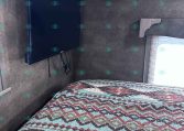 Bedroom in a trailer with a wall-mounted flat-screen TV on the left wall and a multicolored zigzag quilt on the bed.