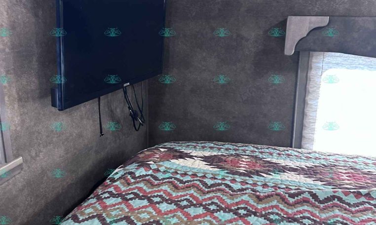 Bedroom in a trailer with a wall-mounted flat-screen TV on the left wall and a multicolored zigzag quilt on the bed.