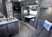 Interior of a modern RV showing a kitchen island with double sink, gray cabinets, and a dinette with bench seating beside a sofa area with a cushion.