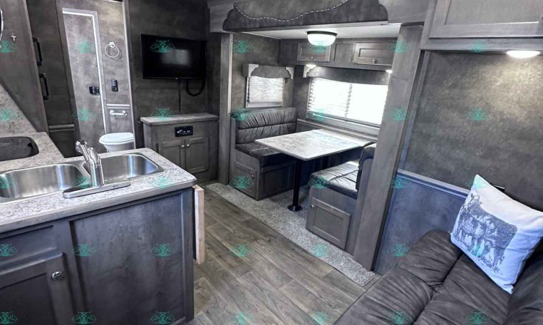 Interior of a modern RV showing a kitchen island with double sink, gray cabinets, and a dinette with bench seating beside a sofa area with a cushion.