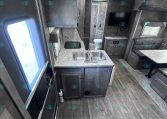 Interior of a modern RV kitchen and living area with sink, cabinets, seating, and a wall-mounted TV.