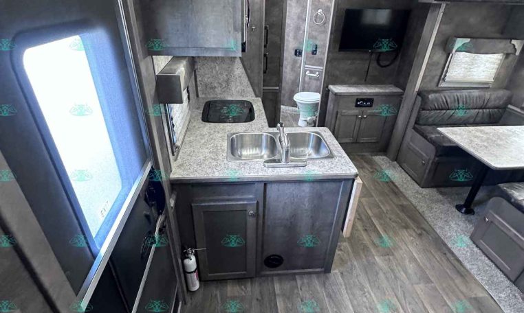 Interior of a modern RV kitchen and living area with sink, cabinets, seating, and a wall-mounted TV.