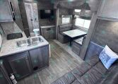 Interior of a modern gray RV with a dual-sink kitchen, counter, and seating area; TV mounted on wall and dinette withbanquette seating by windows.