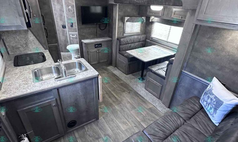 Interior of a modern gray RV with a dual-sink kitchen, counter, and seating area; TV mounted on wall and dinette withbanquette seating by windows.