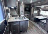 Interior of a travel trailer showing a kitchen with sink, counter, and stove, adjacent to a seating dinette area and mounted TV on the wall