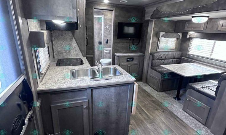 Interior of a travel trailer showing a kitchen with sink, counter, and stove, adjacent to a seating dinette area and mounted TV on the wall