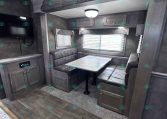 RV interior dining area with booth seating around a marble-pattern table, window blinds, and overhead cabinets; TV mounted on the wall to the left.