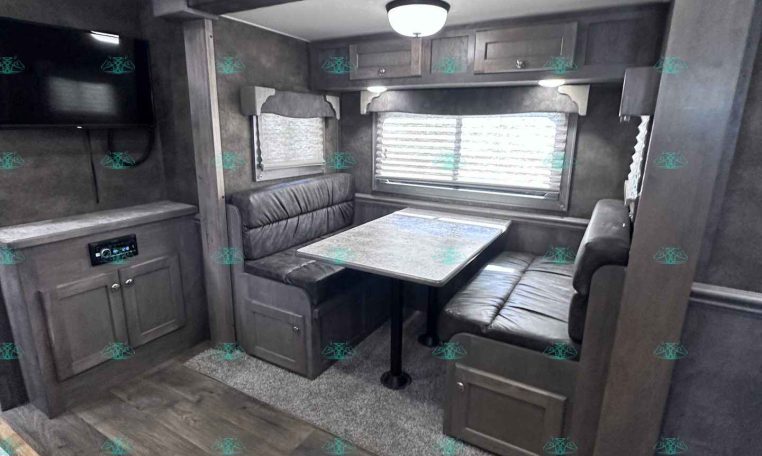RV interior dining area with booth seating around a marble-pattern table, window blinds, and overhead cabinets; TV mounted on the wall to the left.