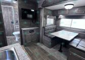 Interior of a gray RV with a kitchenette in the foreground and a dinette booth to the right, multiple cabinets above, and a wall-mounted TV.