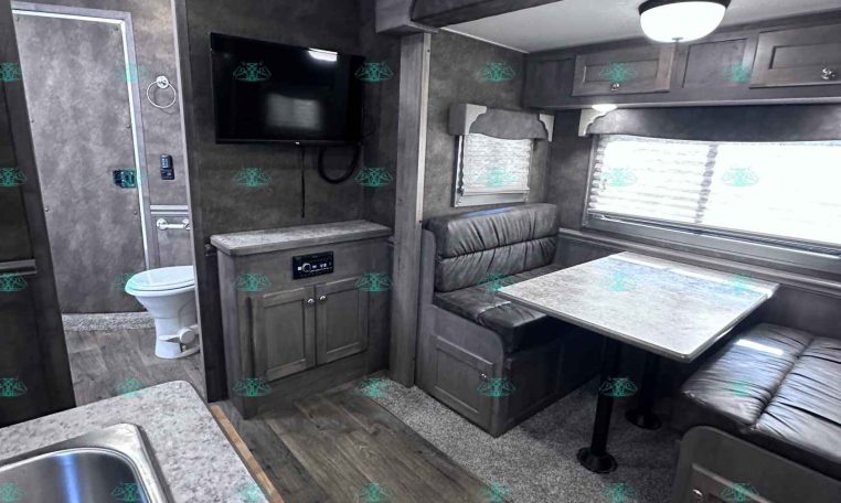 Interior of a gray RV with a kitchenette in the foreground and a dinette booth to the right, multiple cabinets above, and a wall-mounted TV.