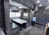 Grey-dominated RV living area with a booth dinette, over-cab bunk, and L-shaped sofa along a corridor of dark wood cabinets and windows.