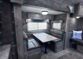 RV dinette with U-shaped gray vinyl bench seating around a rectangular tabletop, string lights above, and cabinets along the wall; windows with blinds in the background.