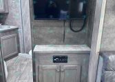 Flat-screen TV mounted on a wood-toned wall above a cabinet in an RV living area, with a countertop and power cables visible.