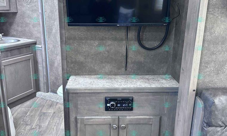 Flat-screen TV mounted on a wood-toned wall above a cabinet in an RV living area, with a countertop and power cables visible.