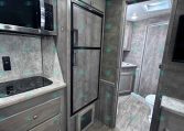 Interior of a modern RV kitchen: stainless microwave above a black stovetop, grey cabinets, full-height fridge, and a doorway leading to a bathroom with a toilet visible.