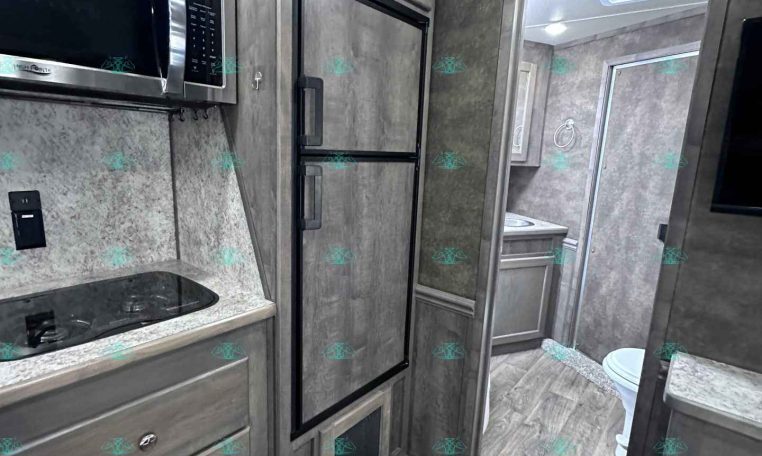 Interior of a modern RV kitchen: stainless microwave above a black stovetop, grey cabinets, full-height fridge, and a doorway leading to a bathroom with a toilet visible.