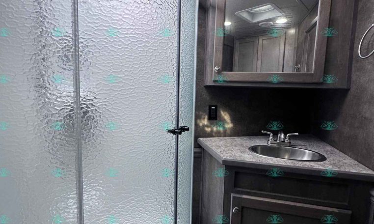 Bathroom inside a trailer: curved glass shower on the left and a dark wood vanity with sink and mirror on the right.