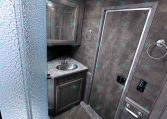 Compact RV bathroom with a frosted glass shower, gray vanity with sink, mirror cabinet, and a toilet in a small trailer interior.
