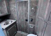 Compact RV bathroom with gray wood vanity, stainless oval sink, mirrored medicine cabinet, towel rings, shower enclosure, and toilet.