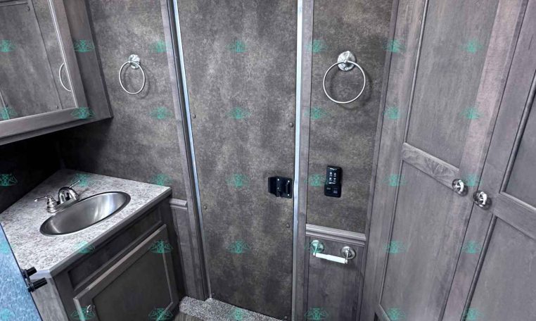 Compact RV bathroom with gray wood vanity, stainless oval sink, mirrored medicine cabinet, towel rings, shower enclosure, and toilet.