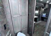 RV bathroom interior with gray wood cabinets, toilet, and doorway to a lounge area with a bunk/sofa visible beyond the door