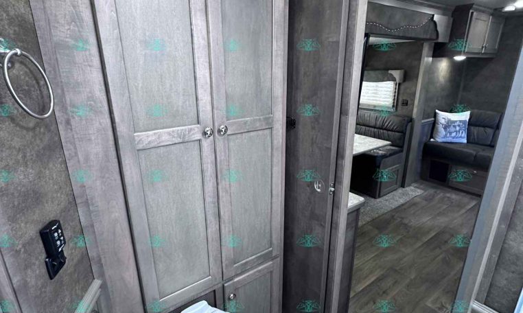 RV bathroom interior with gray wood cabinets, toilet, and doorway to a lounge area with a bunk/sofa visible beyond the door