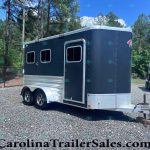Dark blue horse trailer with chrome trim and two windows, parked on a gravel lot beside a white trailer in a wooded area.