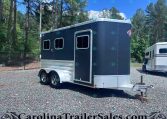Dark blue horse trailer with chrome trim and two windows, parked on a gravel lot beside a white trailer in a wooded area.