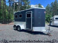 Dark blue horse trailer with chrome trim and two windows, parked on a gravel lot beside a white trailer in a wooded area.