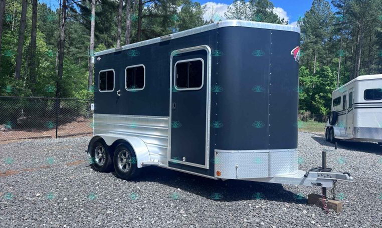 Dark blue horse trailer with chrome trim and two windows, parked on a gravel lot beside a white trailer in a wooded area.