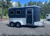Dark blue horse trailer with two windows and chrome trim parked on a gravel lot, trees in the background.