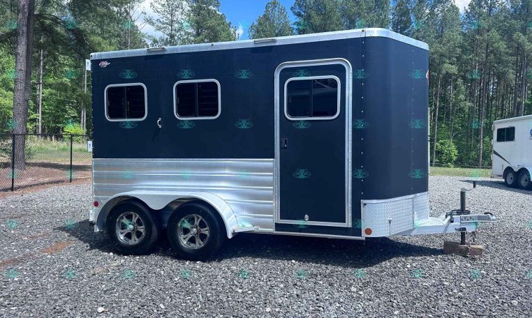 Dark blue horse trailer with two windows and chrome trim parked on a gravel lot, trees in the background.