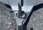 Close-up of a utility trailer hitch with a rusted jack post, safety chains, and a diamond-plate tongue on gravel.