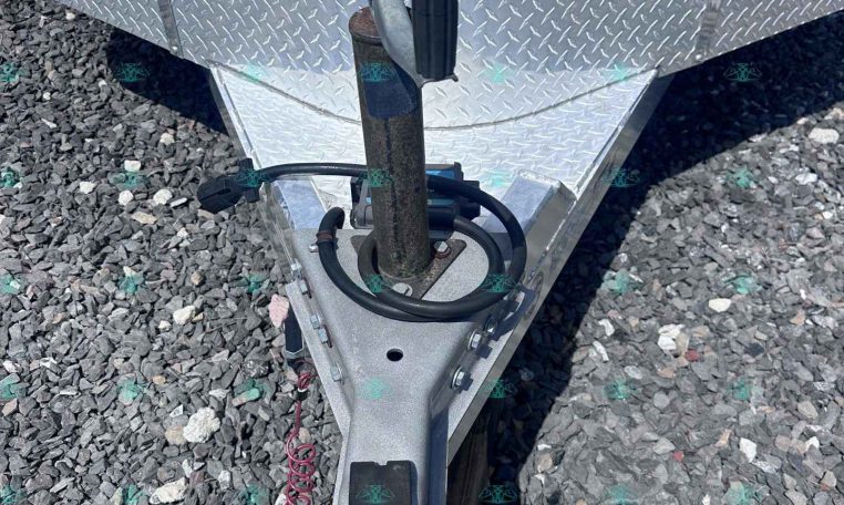 Close-up of a utility trailer hitch with a rusted jack post, safety chains, and a diamond-plate tongue on gravel.