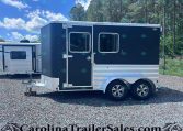 Dark blue enclosed horse trailer with two side-by-side windows and tandem wheels on a gravel lot, trees in the background.