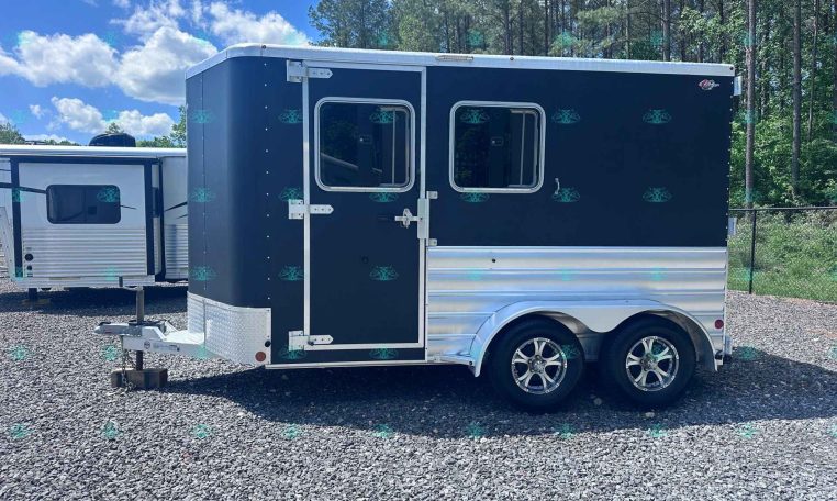 Dark blue enclosed horse trailer with two side-by-side windows and tandem wheels on a gravel lot, trees in the background.