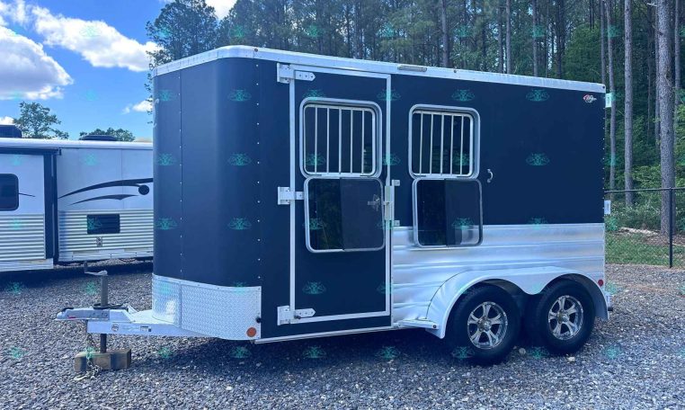 Dark blue horse trailer with two barred windows and aluminum trim, on a gravel lot with trees in the background.