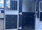Open side door of a dark horse trailer showing an empty interior, parked on gravel with a blue sky above.