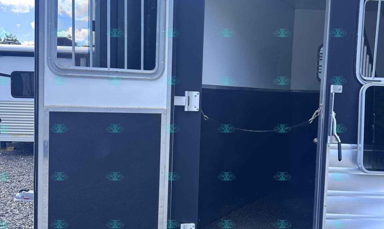Open side door of a dark horse trailer showing an empty interior, parked on gravel with a blue sky above.